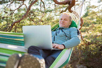 A man working on a laptop while lying in a hammock in the woods. Self-isolation, freelancing, remote work and distancing. Top view of Scandinavian landscape 