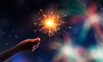 Wall Mural - Hand holding a burning sparkler against fireworks background