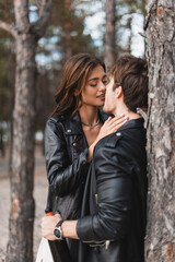 Wall Mural - Selective focus of woman kissing boyfriend in leather jacket with bottle of wine in forest