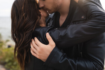 Wall Mural - Man kissing and embracing brunette girlfriend in leather jacket on beach