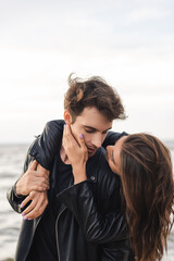 Wall Mural - Young woman touching cheek and embracing boyfriend in leather jacket near sea