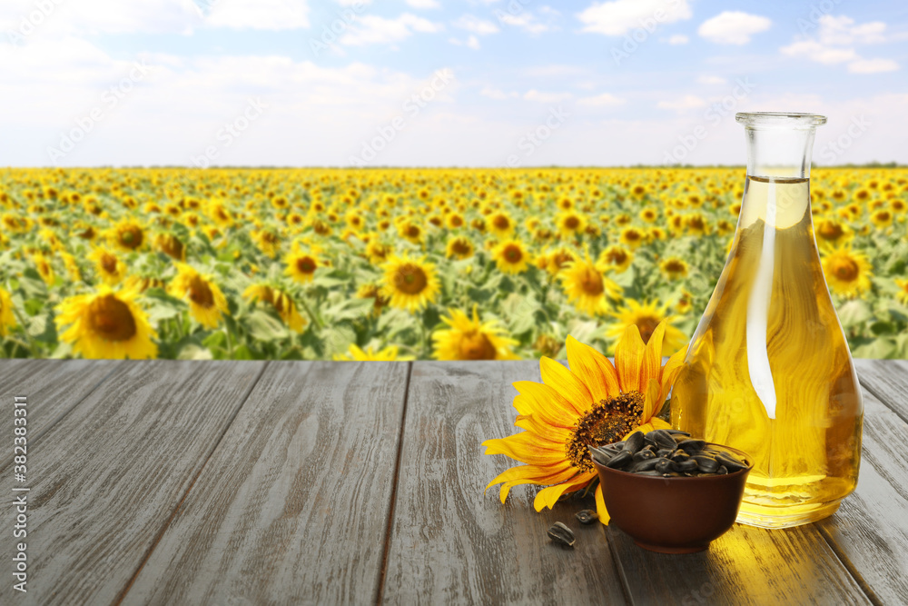 Sunflower oil and seeds on wooden table near blooming field. Space for text - obrazy, fototapety, plakaty 