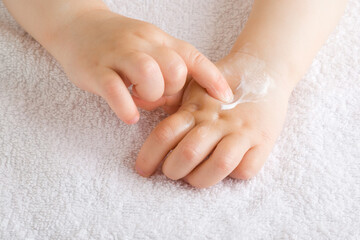 Wall Mural - Baby girl finger applying moisturizing cream on hand on white towel. Care about children clean and soft body skin. Front view. Closeup.