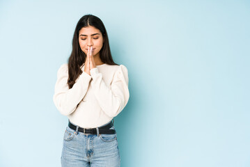 Young indian woman isolated on blue background holding hands in pray near mouth, feels confident.