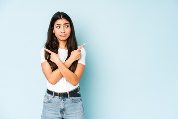 Wall Mural - Young indian woman on blue background points sideways, is trying to choose between two options.