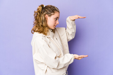 Wall Mural - Young caucasian woman on purple background shocked and amazed holding a copy space between hands.
