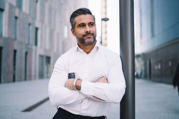 Caucasian male banker with crossed hands standing at urbanity in financial district and thoughtful looking away, pensive businessman thinking about corporate lifestyle pondering in downtown