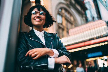 Cheerful pretty hipster girl dressed in trendy outfit enjoying time in urban city standing at street, happy young woman in fashionable leather jacket and glasses smiling during leisure in New York