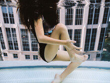 Girl Floating In Sitting Position Underwater In Front Of A Window With A View Of A Large Building. Dreamy.
