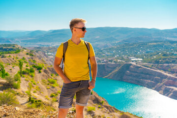 A young man in a yellow shirt is standing above the quarry with blue lake,  quarry is a tourist natural attraction in Crimea