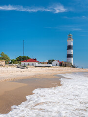 Wall Mural - view to white foam of waves and lighthouse on the beach in summer day with copy space in the sky
