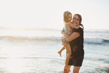 Fun, Energetic Grandma Hugging In Ocean With Young Grandchild - Girl - On Beach At Sunset