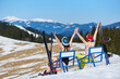 Back view of young couple, sexy woman in bikini with skis and snowboarder man with bare torso sitting in folding chairs with raised arms on background of blue sky and winter snowy mountains panorama.
