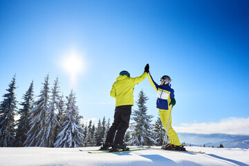 Wall Mural - Couple high-five to each other under sunny blue sky high in mountains. Skiers after successful skiing up to mountain top. Coniferous forest on background. Happiness, success, common goal concept.