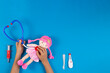 Kid hands play pretend doctor game. Toy stethoscope, pink doll and medicine tools on light blue background. Top view