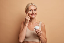 Glad Lively European Woman Of Middle Age Applies Anti Aging Cream On Face Has Natural Beauty Cares About Skin Dressed In Cropped Top Holds Jar Of Cosmetic Product Looks Happily At Camera Poses Indoor