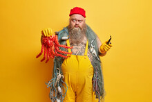 Strict Displeased Bearded Man Seafarer Looks Seriously At Camera Holds Octopus And Smokes Pipe Goes Fishing On Seashore Dressed In Overalls Stands Against Yellow Background. Sailor Style Occupation