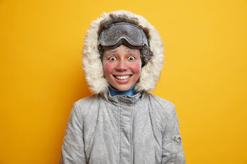 Glad ethnic woman with red frosty cheeks smiles happily feels cold dressed in warm jacket against yellow background goes skiing in mountains during icy snowy weather low temperature and chilling