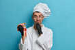Photo of thoughtful male chef looks at crayfish and thinks what to cook for restaurant visitors wears white uniform rubber gloves cooks gourmet dish isolated on blue background. Culinary concept