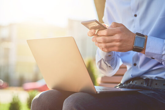 Online business. Cropped shot of a businessman using smartphone and laptop while sitting on the bench outdoors and working remotely