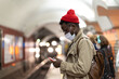 Black millennial man in trench coat, red hat wearing face mask as protection against  covid-19, flu virus, waiting for the train at subway station, using mobile phone. New normal, pandemic concept 
