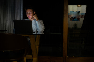 Wall Mural - one young man or teenager working or doing homework at late night in online classes lessons - adult in a table looking and using the laptop smiling and having fun