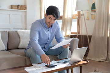 Paying bills. Pensive millennial man involved in paperwork at home, young male sitting on sofa with laptop and calculator preparing financial estimates for loan credit payment, counting rental charge