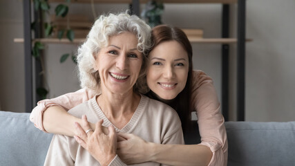 Wall Mural - Head shot portrait young woman hugging smiling mature mother from back, sitting on cozy couch at home, happy beautiful elderly female and adult daughter looking at camera, posing for family photo