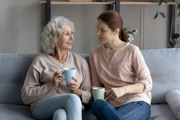 Wall Mural - Happy mature woman with adult daughter drinking tea or coffee, sitting on cozy couch in living room, smiling elderly mother or grandmother chatting with granddaughter, enjoying leisure time