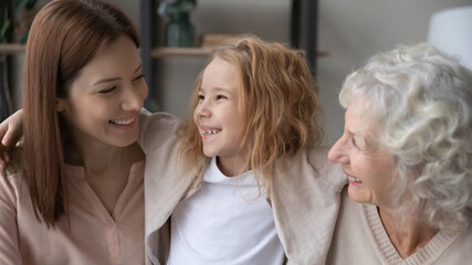 Wall Mural - Close up pretty little girl having fun with young mother and mature grandmother, hugging, cuddling, enjoying tender moment, three generations of women enjoying leisure time together at home