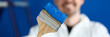 © H_Ko - Close-up of paintbrush covered with blue paint. Smiling worker in uniform on background. Professional interior designer. Renovation and construction site concept