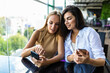 © F8  \ Suport Ukraine - Two cute women in casual clothes are looking at the screen of a mobile phone while sitting in a street cafe. Blogers are reading news on a smart-phone while having coffee break.