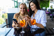 © F8  \ Suport Ukraine - Two women smiling and using cellphones while drinking cocktails at table in cafe