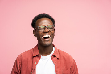 Laughing positive young Afro American man in red shirt in good mood, poses at camera with closed eyes. Overjoyed Black male in spectacles shows her broad smile, isolated on studio pink background. 