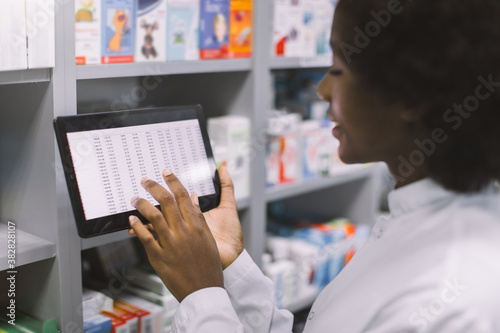 Close-up cropped shot of African American woman doctor or clinical pharmacist using digital tablet while standing in interior of modern hospital pharmacy, making drug quality control