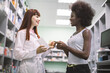 © sofiko14 - Medicine, pharmaceutical care and people concept. Young Caucasian woman apothecary with medicines in hand, counsulting pretty African female customer at pharmacy