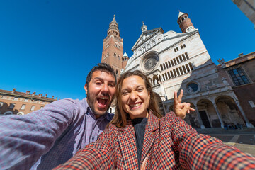 Wall Mural - happy tourist couple visiting the city of Cremona taking selfie in the middle of main square of Cremona with Cathedral, baptistery and Torrazzo bell tower . 