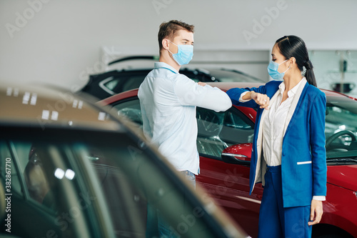 Sales manager and customer in medical masks doing elbow bump when greeting each other in car dealership