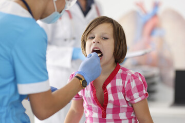 Wall Mural - Doctor in blue suit look at child throat with wooden stick. Child in plaid shirt sit at doctor office and opened her mouth wide.