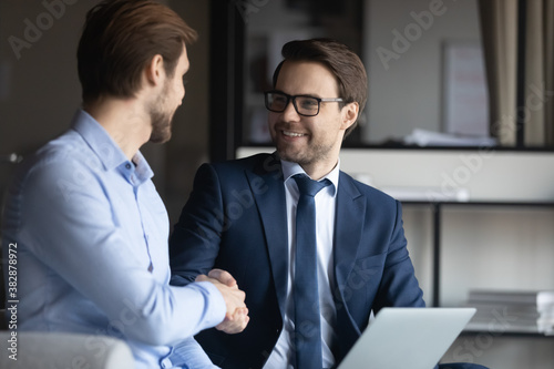 Smiling businessman in eyeglasses and formal suit shaking hands with partner, establishing cooperation. Happy executive manager thanking young employee for good job, satisfied with project results.