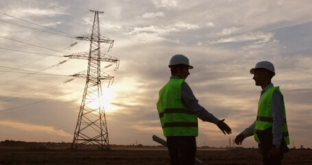 Wall Mural - two engineers in white safety helmets and green signal vests shake hands in front of a high-voltage 