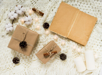 Winter composition with an old book, gifts, pine cones, cinnamon, a cotton branch and glowing garlands on a light knitted blanket. Flat lay, top view. Warm cozy Christmas.