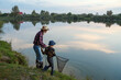 Cute 10-aged boy with his experienced grey-bearded 70-aged grandpa catching fish on the lake with landing net at sunset