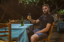 Young Man Is Holding Glass Of Beer In Ambient Of Traditional Greek Tavern Or Restaurant At Night While Looking From Camera. Table With Checkered Table Cloth With Beer Bottle And Wooden Chairs Visible.