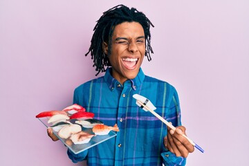 Young african american man eating sushi using chopsticks winking looking at the camera with sexy expression, cheerful and happy face.