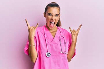 Young hispanic woman wearing doctor uniform and stethoscope shouting with crazy expression doing rock symbol with hands up. music star. heavy concept.