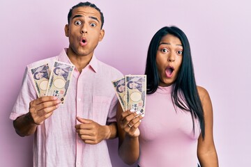 Canvas Print - Young latin couple holding japanese yen banknotes scared and amazed with open mouth for surprise, disbelief face