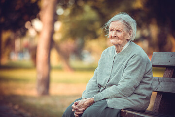 Portrait of senior woman sitting on bench in autumn park. Old lady feeling lonely and sad.