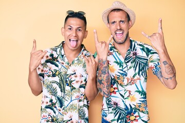 Young gay couple of two men wearing summer hat and hawaiian shirt shouting with crazy expression doing rock symbol with hands up. music star. heavy music concept.