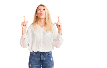 Young beautiful blonde woman wearing casual shirt amazed and surprised looking up and pointing with fingers and raised arms.